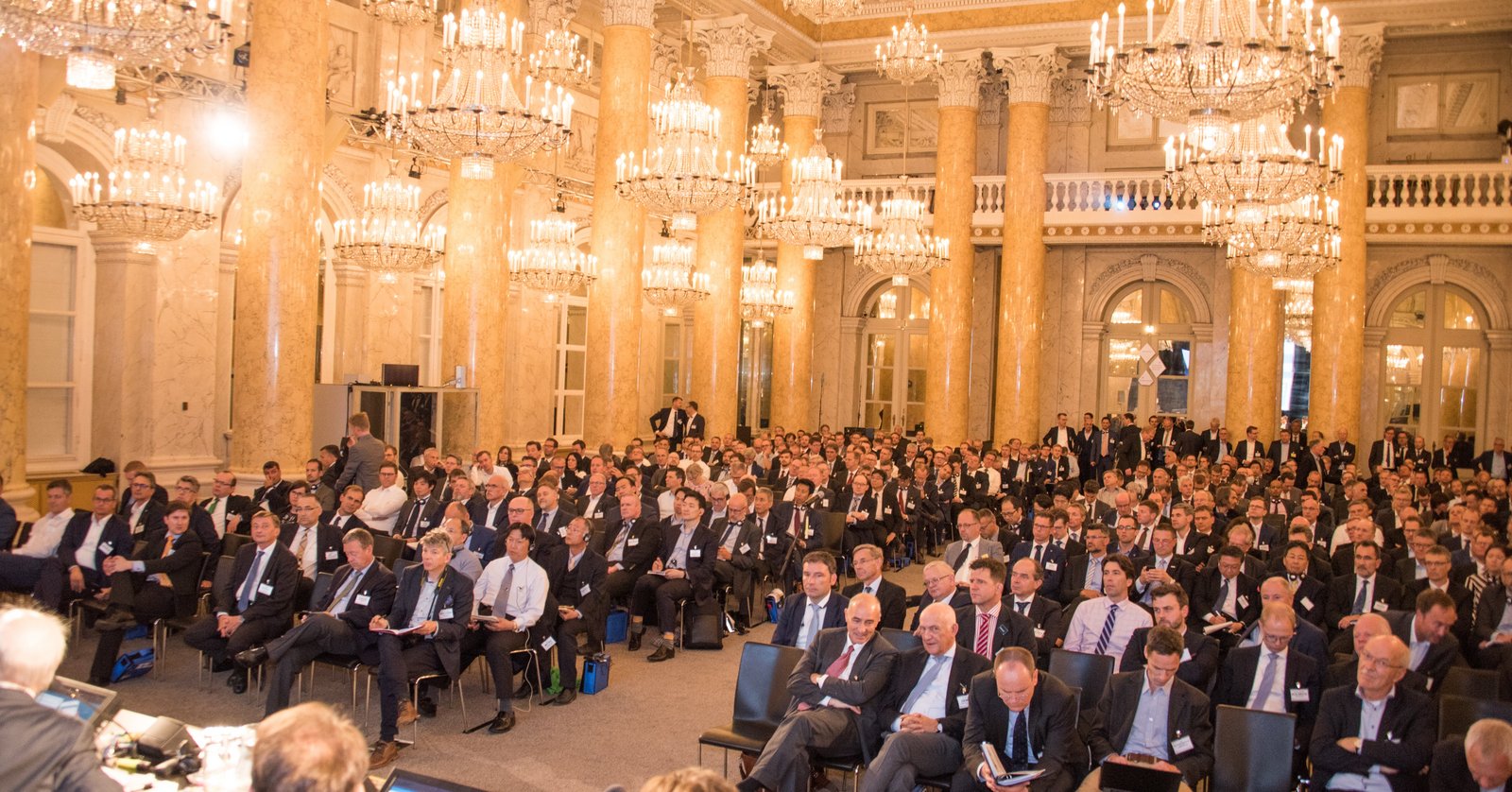 Besucher im Anzug sitzen im Saal mit leuchtenden Kronleuchtern
